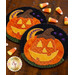 Two wool coasters shaped like Halloween pumpkins, featuring a smiling face and a brown stem, are placed on a wooden surface alongside pieces of candy corn. The coasters have colorful stitching details and a green base.