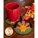 A red mug sits on two round coasters featuring a turkey design, surrounded by autumn leaves and berries on a wooden table.