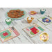 A table set with colorful, patterned table mats featuring festive designs, a plate of cookies, small cups with marshmallows, and a bowl of assorted cookies. The scene has a rustic wooden background.