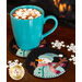 A turquoise mug filled with hot chocolate topped with marshmallows sits on a round black coaster featuring a smiling snowman. Snowflakes are scattered around the table, and a fireplace can be faintly seen in the background.