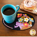 A turquoise coffee mug sits on a decorative black wool coaster featuring a colorful floral design, next to a wooden plate holding a cinnamon roll and a fork.