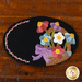 An oval wool mat with a black background featuring a colorful arrangement of flowers in a basket, accented by a purple bow. A decorative border made of small pink flowers runs along one edge.