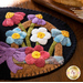 A decorative mat featuring a variety of colorful wool flowers, including purple, pink, white, and blue, arranged in a basket-like design against a black background.