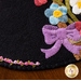 A close-up of a wool mat featuring colorful wool flowers and a purple bow on a black background, with an embroidered rose edge detailed in pink and green stitching.