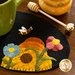 A circular black placemat featuring colorful felt appliqués of flowers, a beehive, and a bee. A green mug is in one corner, and a honey dipper is resting nearby on a wooden table.