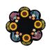 A decorative round wool mat featuring sunflowers, colorful flowers, and watering can illustrations arranged around a central black space. The design has a flower-like silhouette.