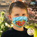 A young boy wearing a colorful face mask with the word HELLO in a comic book style. He has short brown hair and is standing outdoors with greenery in the background.