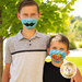 Two boys are standing together outdoors, smiling while wearing colorful face masks. The older boy on the left has a light blue mask with a printed mustache, and the younger boy on the right is wearing a mask featuring the word Hello in a comic-style design. They are surrounded by green foliage.