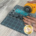 A hand holds an orange rotary cutter, while cutting blue fabric between a ruler and a gray cutting mat.