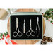 A set of four metallic sewing tools displayed in a black case on a wooden surface, with holiday decor.