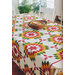 A colorful quilted tablecloth featuring a geometric pattern in red, green, yellow, and white covers a dining table. A wooden bowl sits in the center, with light streaming in through a nearby window, and plants visible in the background.