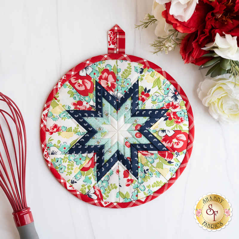 Round folded star hot pad on a white countertop with a bouquet of red and white roses and whisk nearby