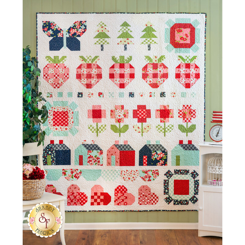 A colorful quilt hanging on a wall features various patchwork designs, including butterflies, trees, strawberries, flowers, and houses, in a combination of red, blue, green, and white fabrics. The quilt has a cheerful, whimsical aesthetic, with bold patterns and vibrant colors.