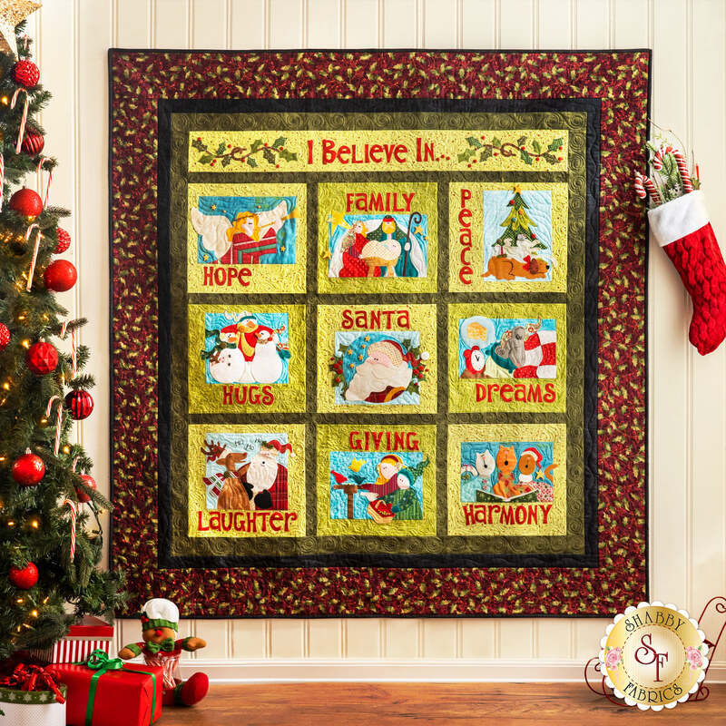 A decorative quilt hanging on a wall, featuring a grid of eight colorful squares with various holiday-themed illustrations and words: Hope, Family, Peace, Santa, Hugs, Giving, Laughter, and Harmony. The quilt is bordered by a floral design and is surrounded by a Christmas tree and a festive stocking.