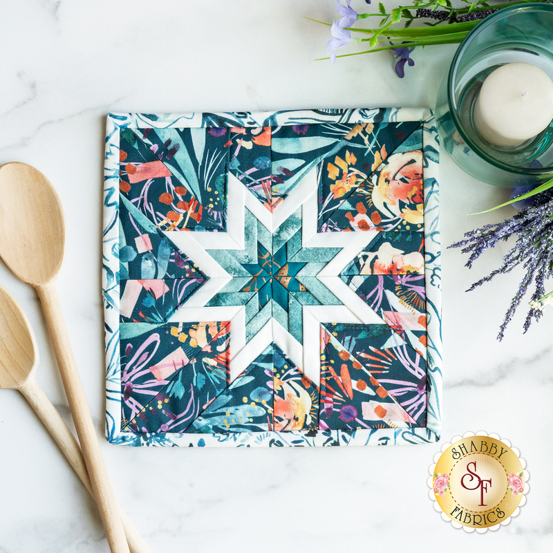 A light teal square hot pad on a white marble countertop with wooden spoons and a candle