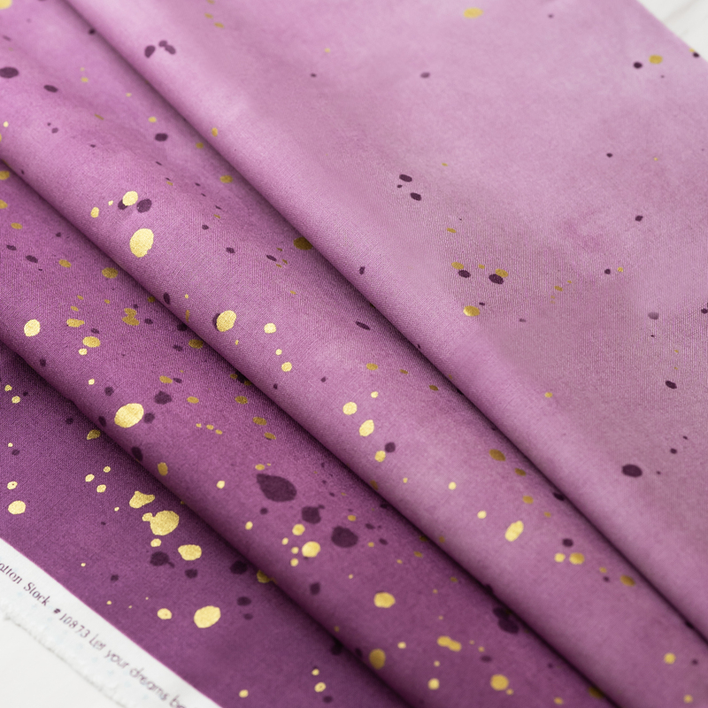 A close-up of folded fabric featuring a gradient of purple shades with scattered gold and dark purple speckles.