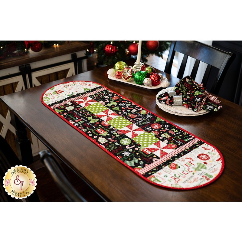 Christmas themed table runner featuring phrases, ornaments, and snowflakes, designed with rounded sides and central pinwheels.