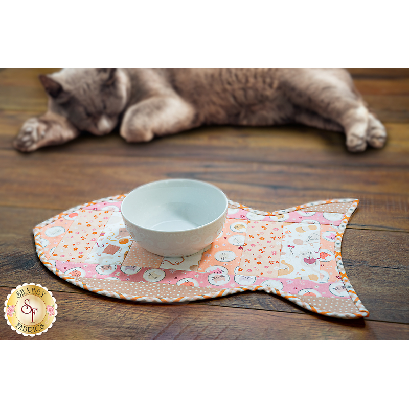 Fish shaped cat pet placemat made of strips of pink and cream cat themed fabrics holding a white bowl near a resting cat.