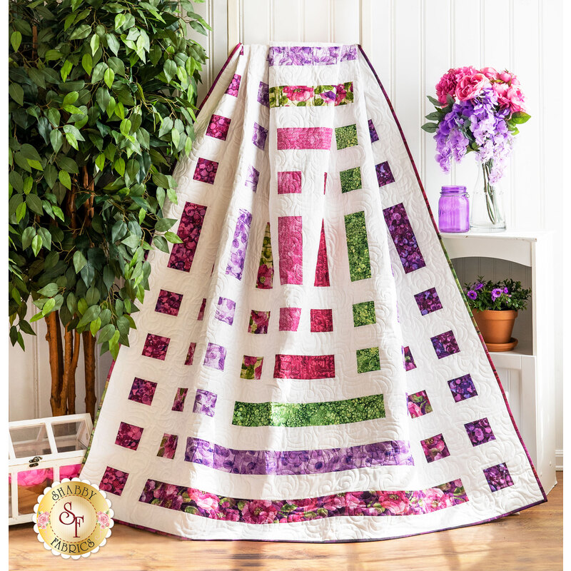 Quilt of squares and rectangles made of pink, purple, and green floral fabrics on white background draped over furniture.