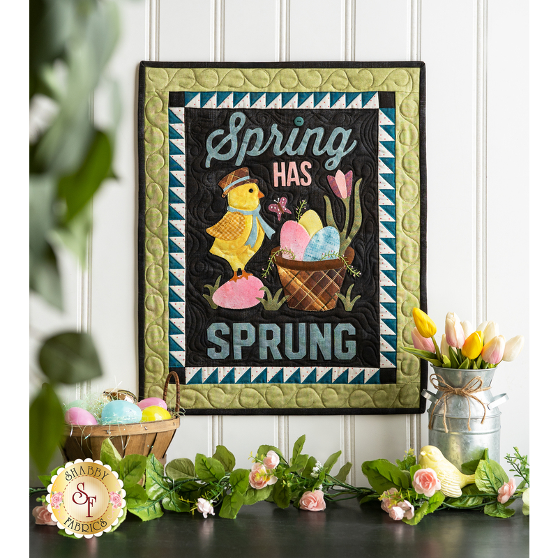 Wall hanging featuring a bird with scarf and hat next to a basket of colored eggs on black with the phrase 