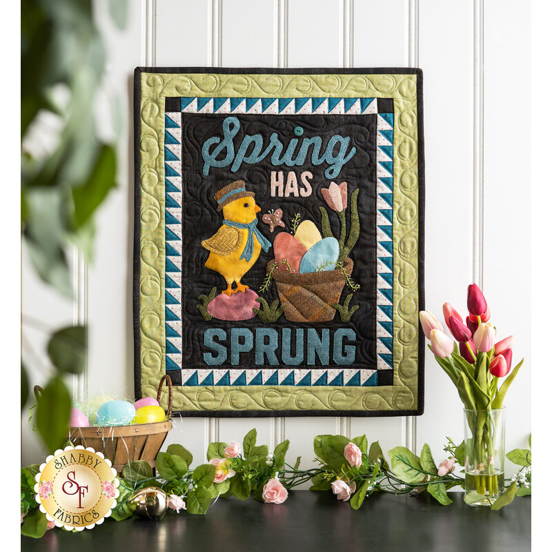 Wall hanging featuring a bird with scarf and hat next to a basket of colored eggs on black with the phrase 