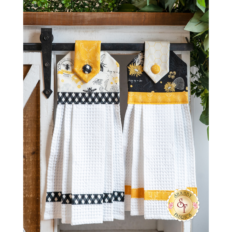 Hanging towel made of white bee themed fabrics with black and yellow accents next to matching towel in black.