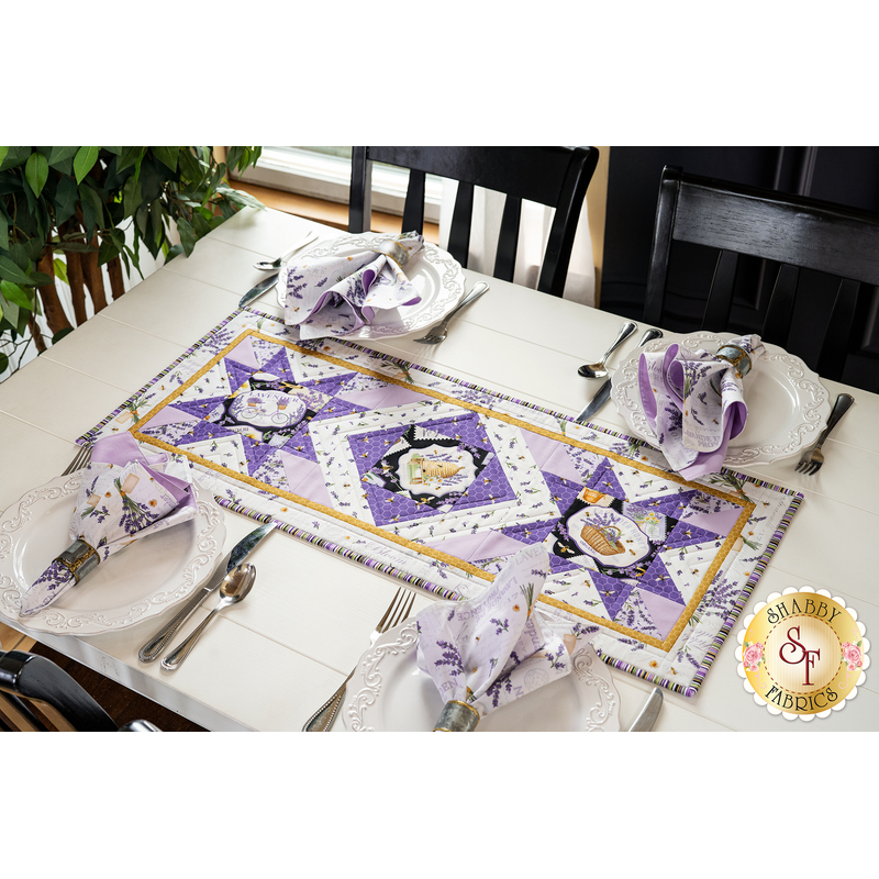 Table runner with geometric design with white and purple lavender and bee themed fabrics on table set with matching napkins.