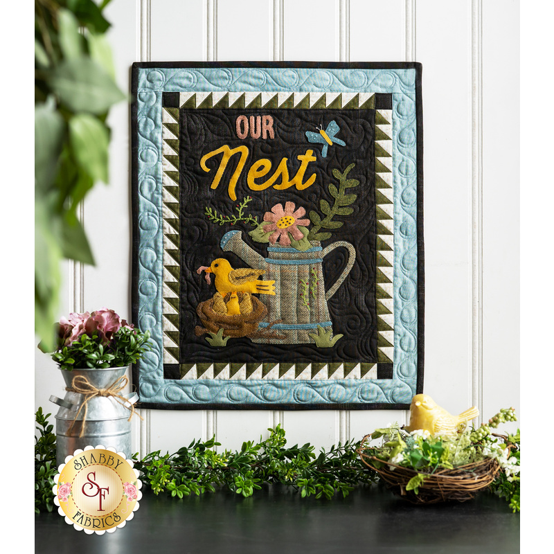 Wall hanging featuring appliqué of a nest of birds next to a watering can full of flowers and the phrase 