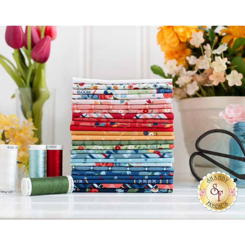 A stack of neatly folded fabric in various colors and patterns sits on a table, surrounded by spools of thread and flowers. The fabrics feature floral designs, stripes, and solid colors, creating a vibrant and organized display.