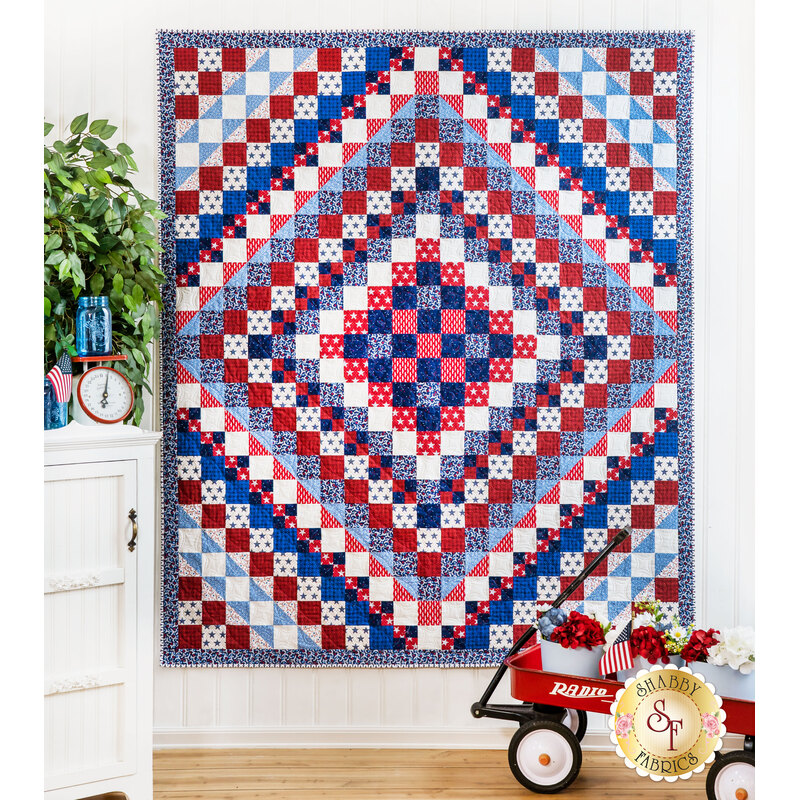 A colorful quilt in red, blue, and white showcases a diamond pattern, displayed on a wall.