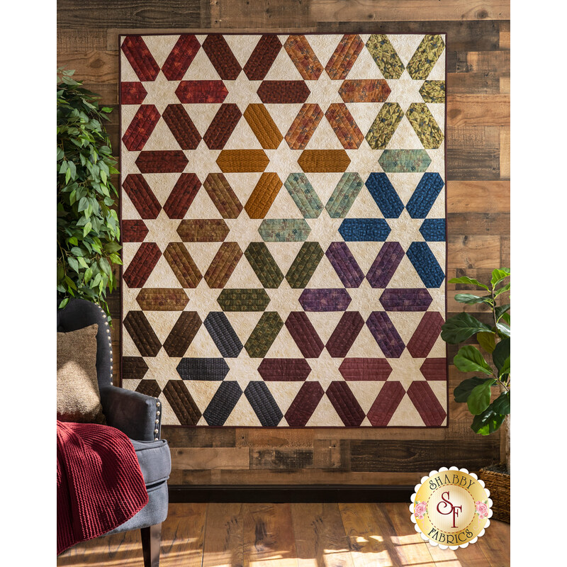 A colorful quilt with star patterns hangs on a wooden wall next to a cozy chair and greenery.