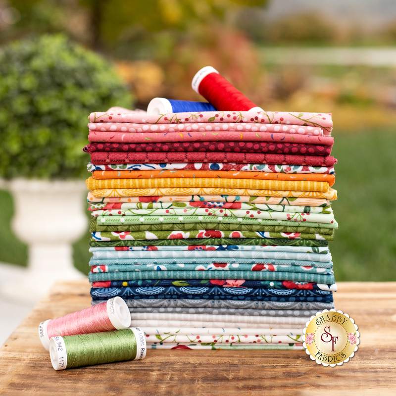 A neatly stacked collection of colorful fabrics in assorted patterns, with three spools of thread placed in front. The background features a blurred garden setting.