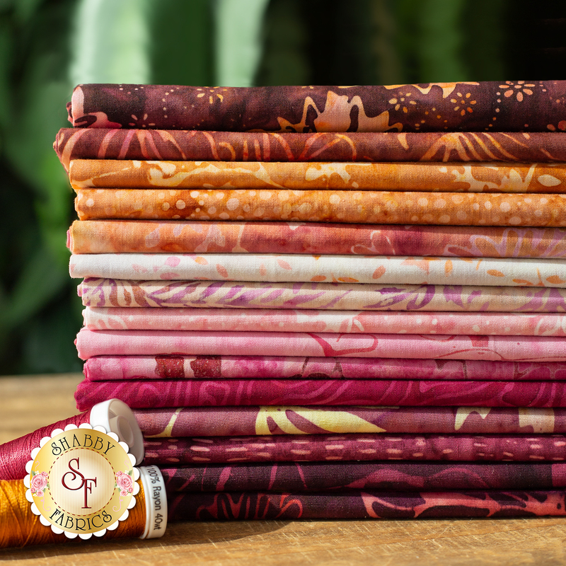 Close up photo of a stack of batik fabric arranged in a gradient of brown and pink with two spools of thread on the counter and greenery in the background.