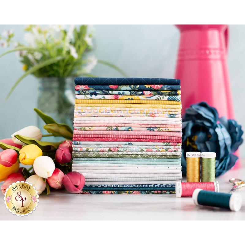 A stack of neatly arranged colorful fabric rolls sits on a table next to a bouquet of tulips and spools of thread. In the background, there is a vase with green leaves and a large blue flower.