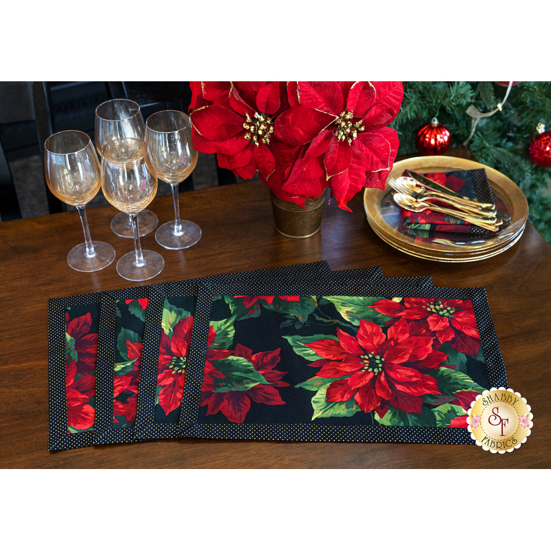 4 Christmas themed poinsettia placemats laid flat on a wood table with other Christmas decor