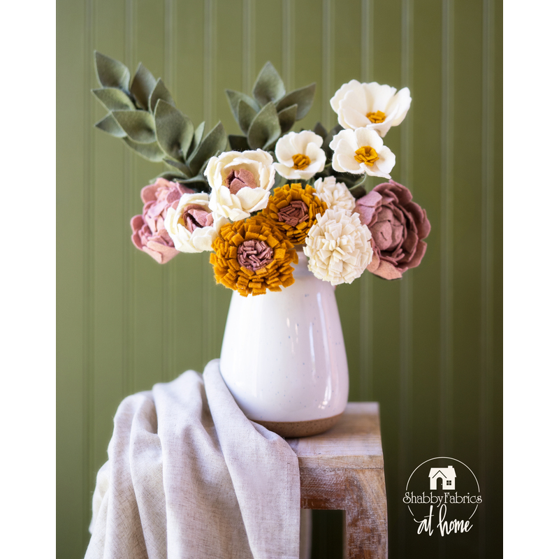 Flowers made of fabric being displayed in a white vase on small wood table.