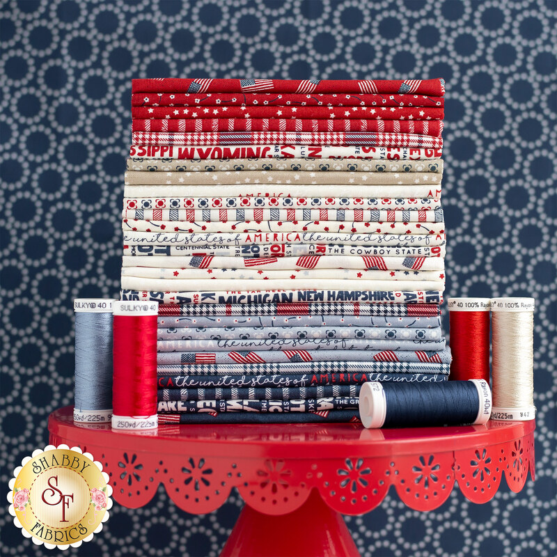 A stack of neatly arranged fabrics in red, white, and blue colors, featuring various patterns and text that represent different U.S. states. The fabrics are displayed on a round red table with a decorative edge, next to several spools of thread in coordinating colors. The background has a subtle pattern.