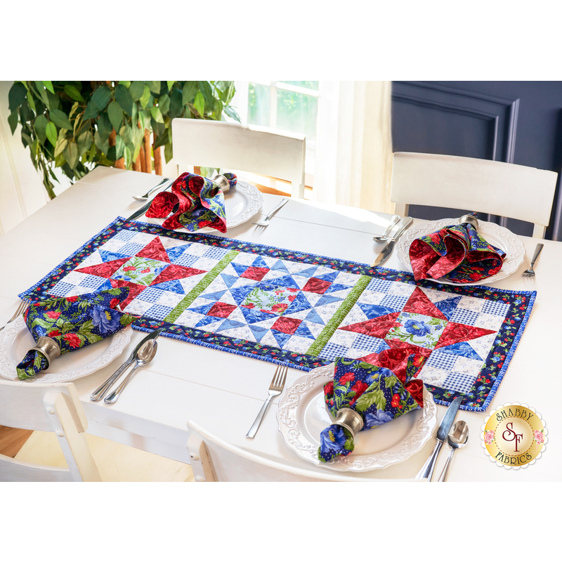 A red, white, and blue table runner made with floral fabrics laid flat on a white table next to cloth napkins