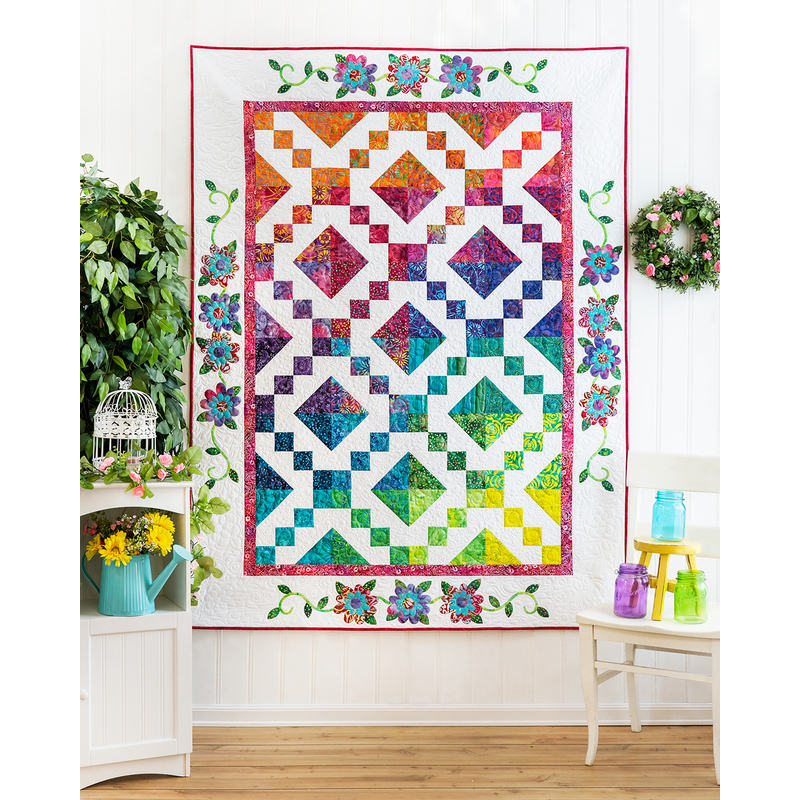 A colorful quilt featuring diamond and square patterns, displayed against a white wall with foliage.