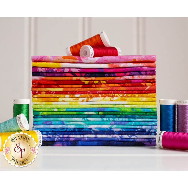 A stack of colorful fabric bundles arranged in order from top to bottom in shades of pink, orange, yellow, green, blue, and purple, with several spools of thread in coordinating colors placed beside the stack. A logo for Shabby Fabrics appears in the bottom left corner.