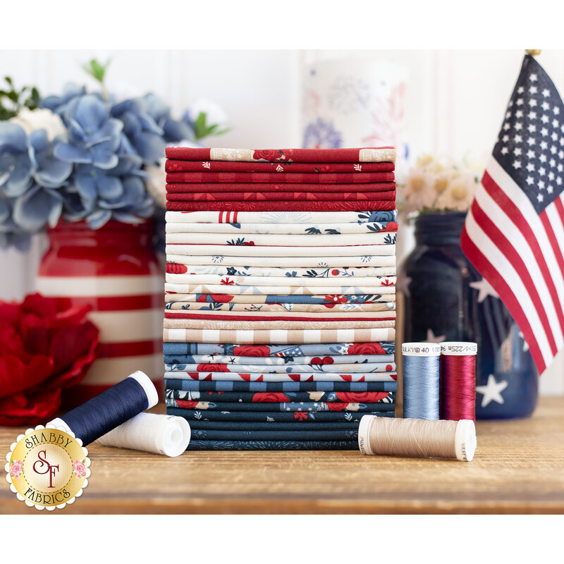 A neatly arranged stack of fabric squares in red, white, blue, and cream colors, featuring various patterns. Spools of thread in coordinating colors are placed in front, alongside a small American flag and decorative flowers.