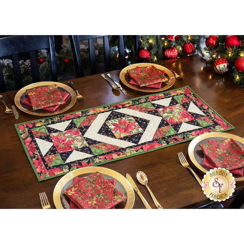 Photo of the Madison table runner made with Christmas fabrics with gold accents on a wooden table with four place settings with matching napkins and a decorated Christmas tree on one side.