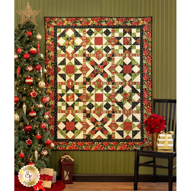 A festive quilt with geometric patterns hangs on a green wall, next to a decorated Christmas tree.