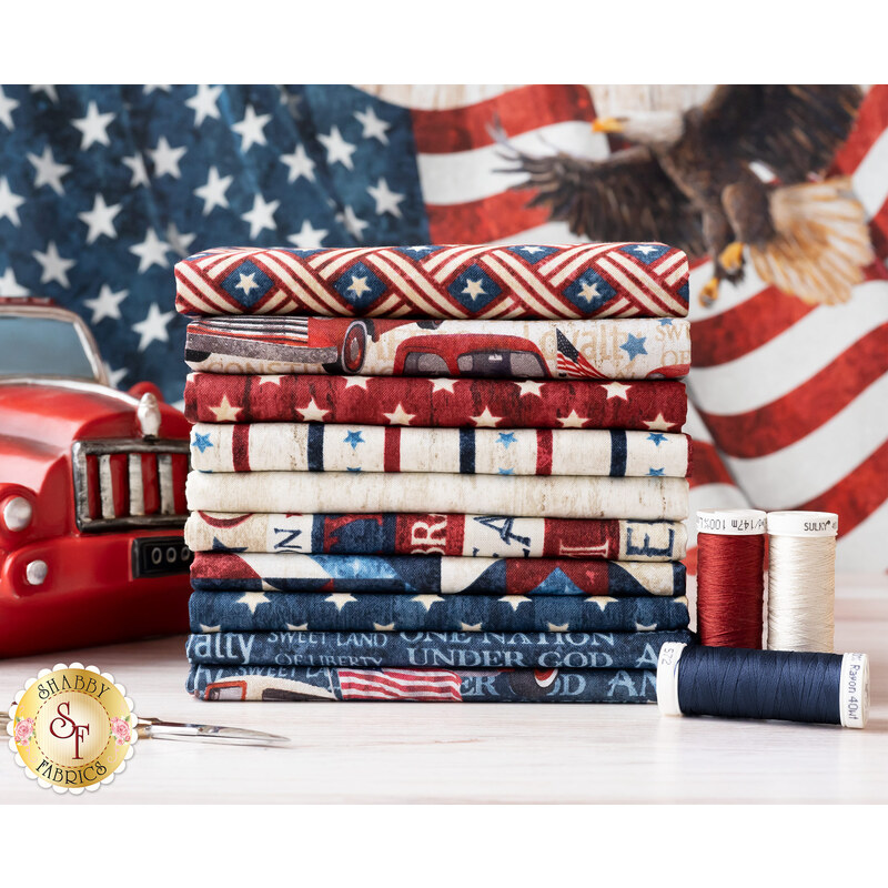 A stack of various patriotic fabric squares in red, white, and blue, featuring patterns like stripes, stars, and text. In the foreground, there is a pair of scissors and several spools of thread in matching colors. A vintage red car is partially visible on the left, with an American flag design in the background.