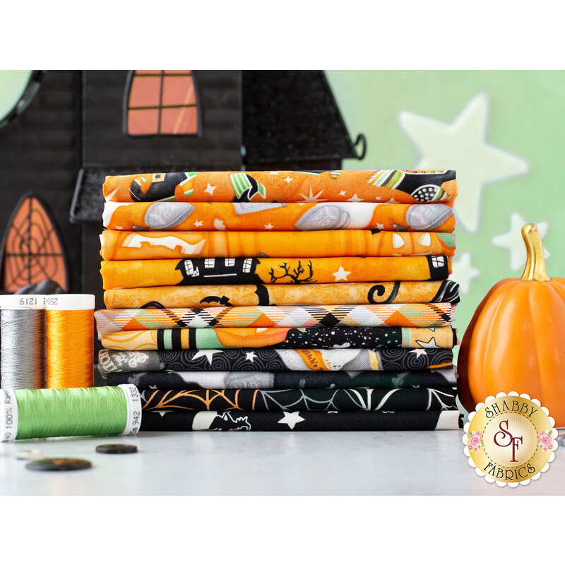 A stack of colorful Halloween-themed fabrics in orange and black, with a pumpkin and spools of thread in the foreground. A black haunted house is partially visible in the background.