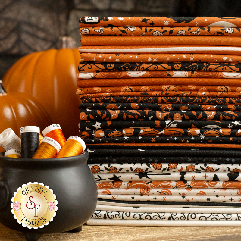 A stack of assorted Halloween-themed fabrics in orange, black, and white, with patterns including pumpkins and stars. There are spools of thread in shades of orange and white next to a decorative black cauldron. A pumpkin is also visible in the background.