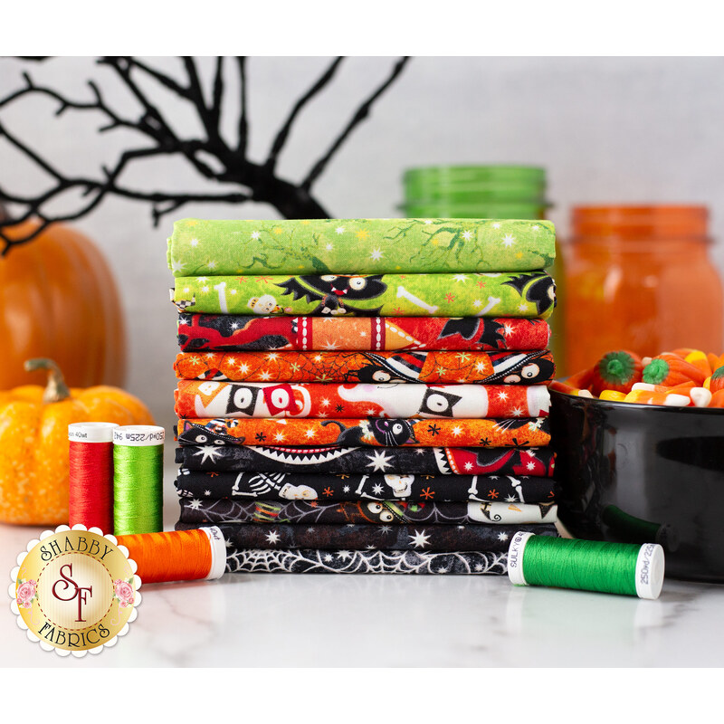 A stack of vibrant Halloween-themed fabric bundles in various colors, including green, orange, black, and white, is displayed alongside spools of thread in green, orange, and red. In the background, there are decorative pumpkins and jars filled with candy.