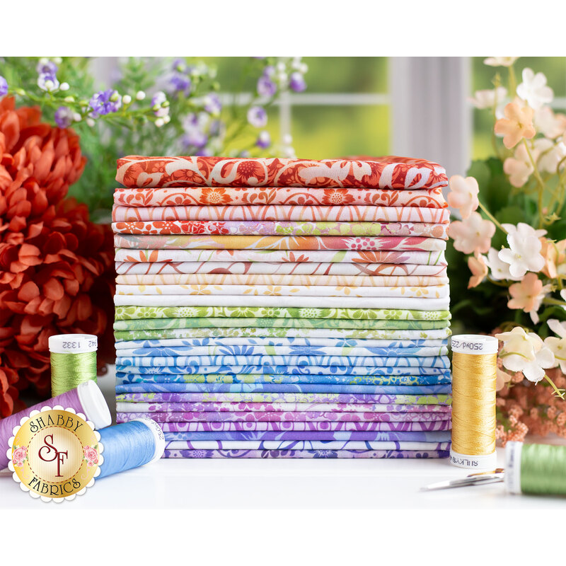 A stack of colorful patterned fabric bolts arranged in a gradient from red to purple, with spools of green and yellow thread beside them, set against a backdrop of flowers and greenery.