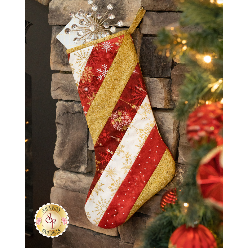 A festive Christmas stocking with diagonal red, white, and gold stripes adorned with snowflake patterns, hanging by a stone fireplace, surrounded by holiday decorations.