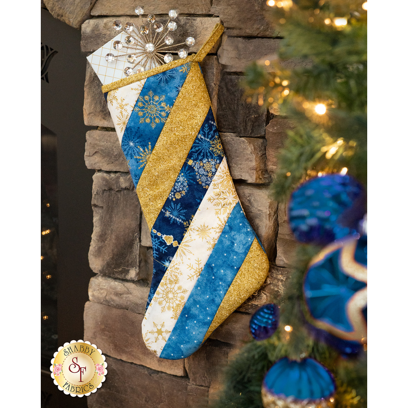 A decorative holiday stocking hangs from a stone fireplace, featuring blue, white, and gold stripes with snowflake designs. A silver snowflake ornament decorates the top of the stocking. Christmas ornaments are visible in the surrounding greenery.